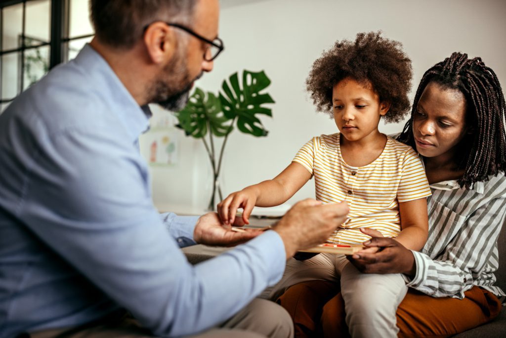 A young African American girl is handed a tablet from a psychiatrist while she sits on her mother's lap