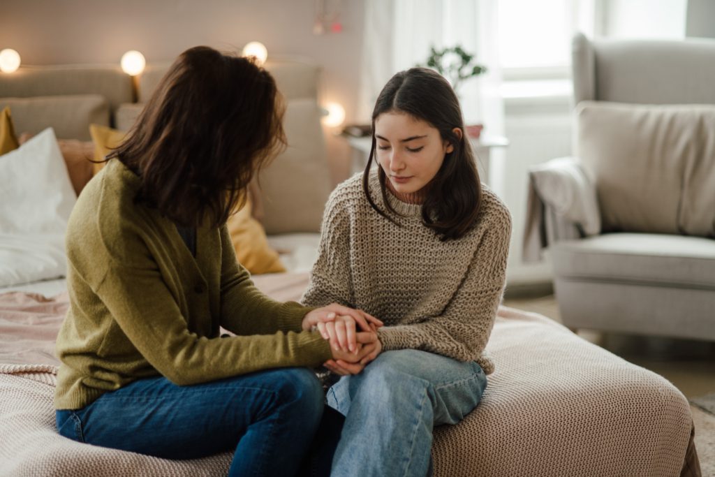 A mother comforts her teen daughter as they sit on the edge of the girl's bed