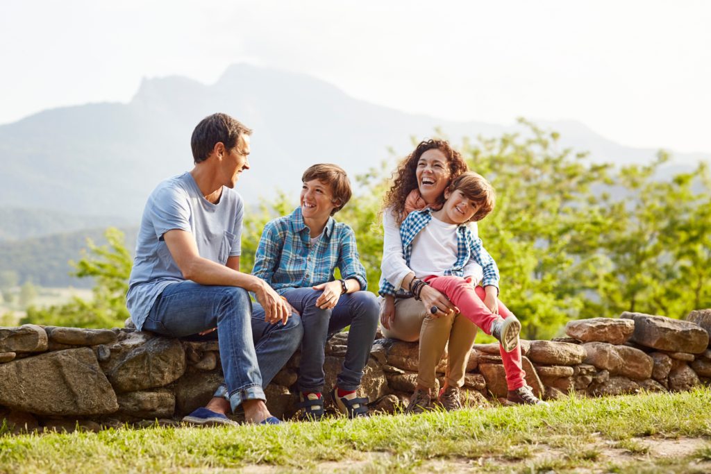 A happy family sits on a small stone wall while taking a hike on a sunny day