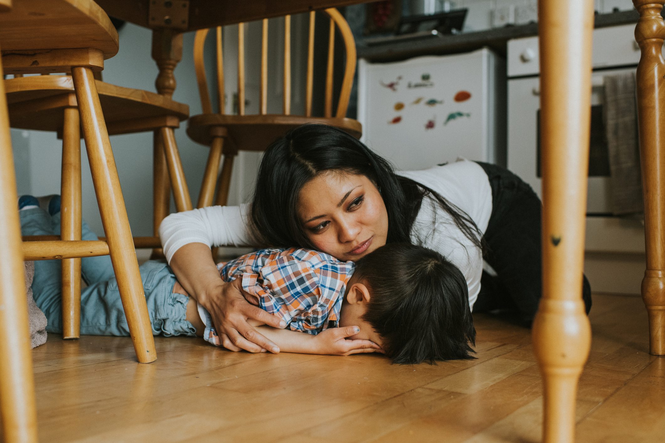 An upset little boy lies face down on the floor, under a kitchen table, comforted by his mother